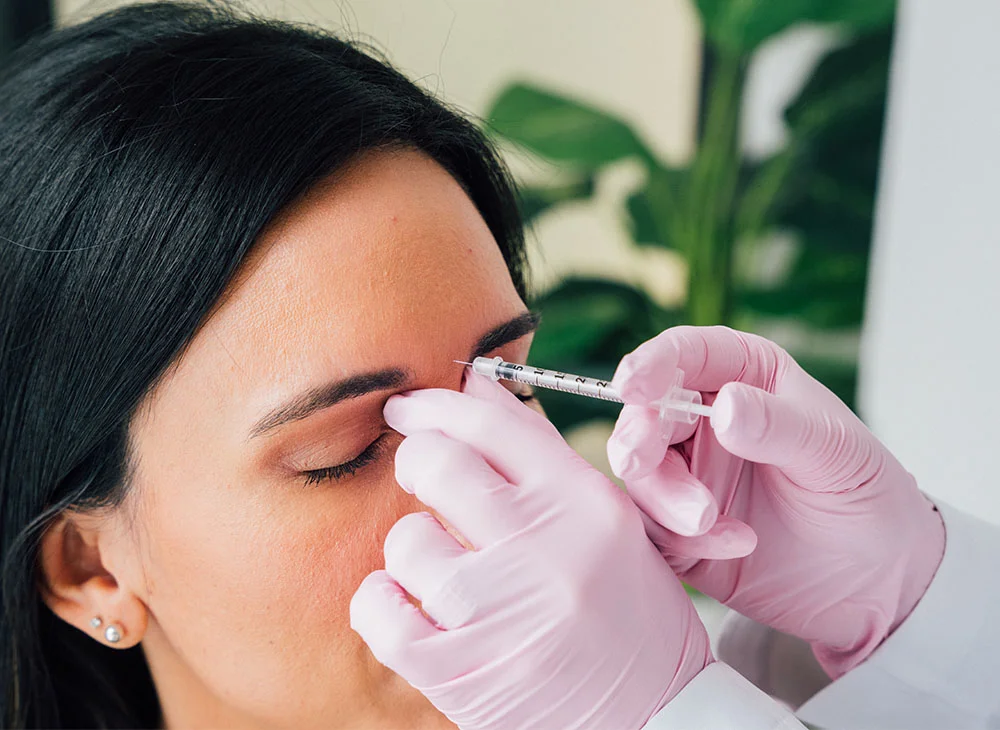 Close-up of a woman receiving a cosmetic injection, likely Botox, in a serene spa setting. The patient, with eyes closed and long dark hair framing her face, relaxes as a medical professional in pink gloves administers a precise injection to her forehead. The presence of plants in the background further enhances the relaxing and rejuvenating atmosphere of the spa. - Botox and Nuermodulators in San Clamente, Aliso Viejo and Rancho Santa Margarita, CA