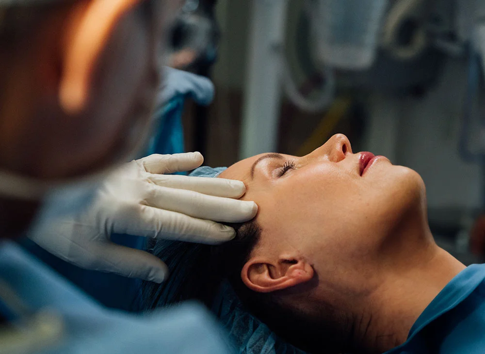 Close-up of a woman lying supine with eyes closed as a medical professional in white gloves gently tends to her forehead. The bright lighting highlights the interaction, with a blurred background showing medical equipment and personnel in blue-gray scrubs. - Brow and Forehead Lift in San Clamente, Aliso Viejo and Rancho Santa Margarita, CA