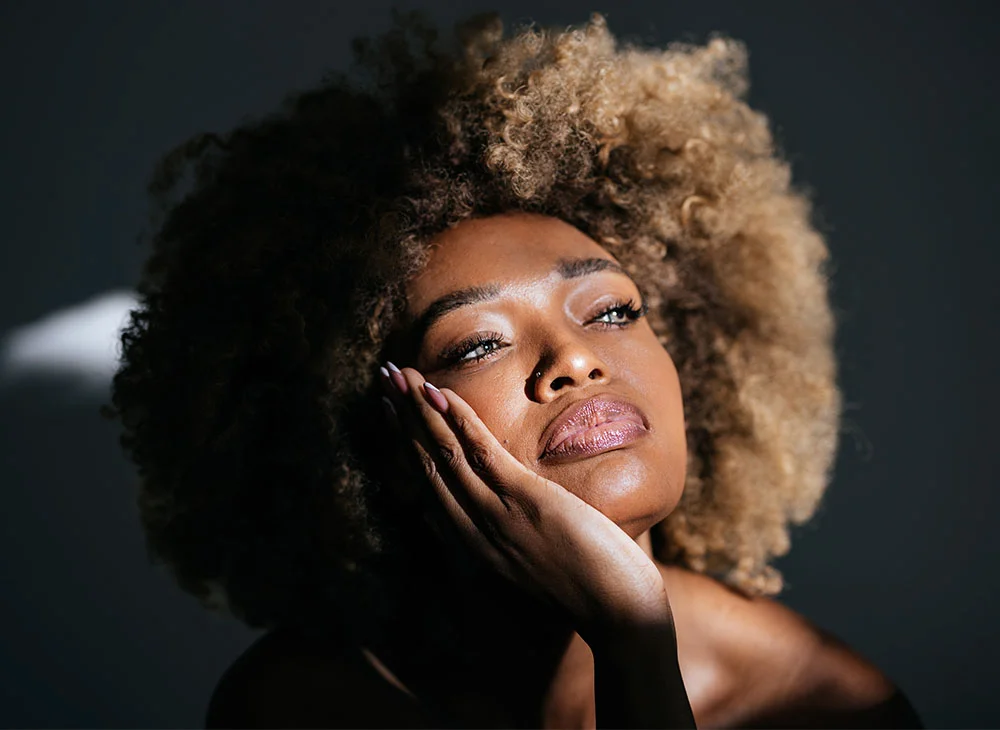 Portrait of a woman with a textured afro hairstyle and a thoughtful expression. The soft light and dark background create a striking image that highlights her natural beauty. - Microeeding in San Clamente, Aliso Viejo and Rancho Santa Margarita, CA