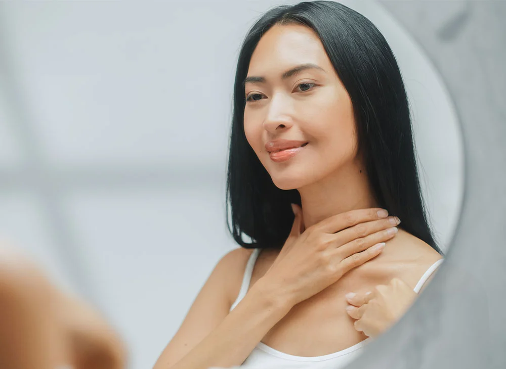 A woman of Asian descent examines her neck and chest in a bathroom mirror. With long dark hair, she gently touches her skin, her expression soft and focused. She wears a white sleeveless top. The neutral-toned background and soft lighting enhance the calm, reflective atmosphere. - Neck Filler in San Clamente, Aliso Viejo and Rancho Santa Margarita, CA