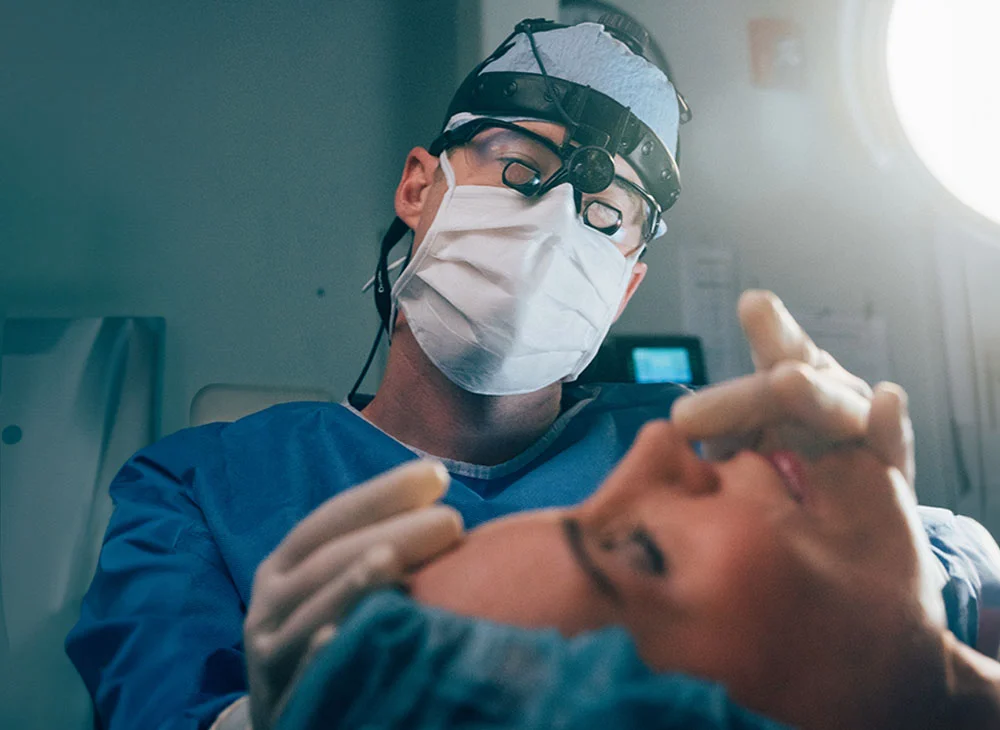 A close-up shot of a surgical procedure being performed by Dr. Brandon. The surgeon, wearing a blue gown, mask, and eyewear, is focused on the patient who is lying face up. Medical instruments and equipment are visible in the background. - Rhinoplasty in San Clamente, Aliso Viejo and Rancho Santa Margarita, CA