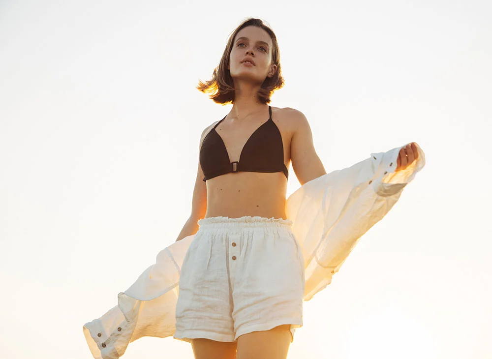 A full-length shot of a young woman with short brown hair standing outdoors. She wears a white linen shirt draped over her shoulders and a bikini top. The light is soft and golden, creating a warm and inviting atmosphere. - Tummy Tuck in San Clamente, Aliso Viejo and Rancho Santa Margarita, CA